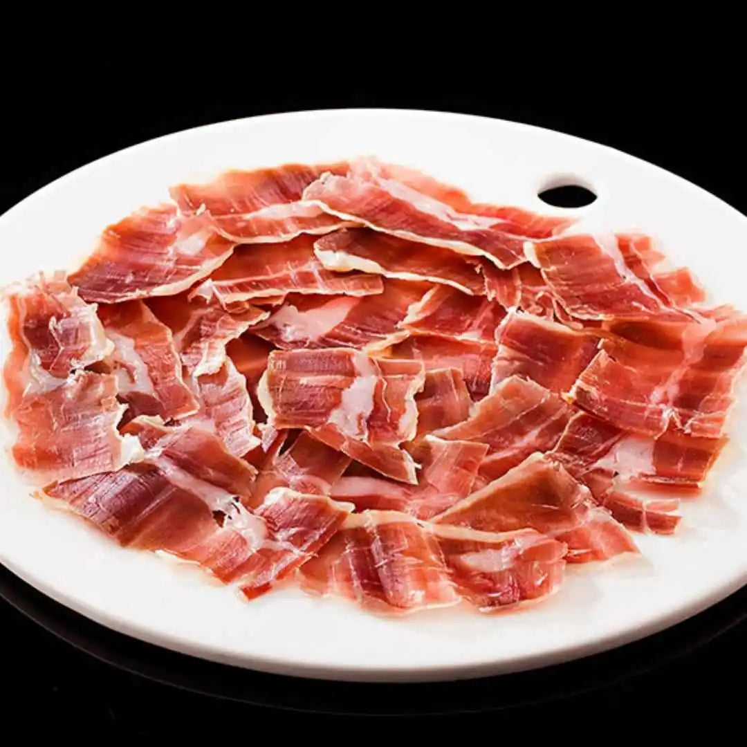 Tranches fines de jambon d’agneau rosé, disposées sur assiette blanche ronde avec fond noir.